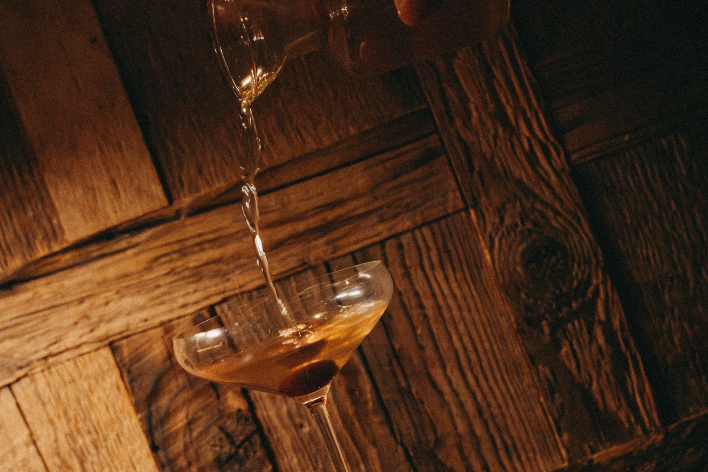 A drink being poured into a coupe glass on a dark wooden bar.